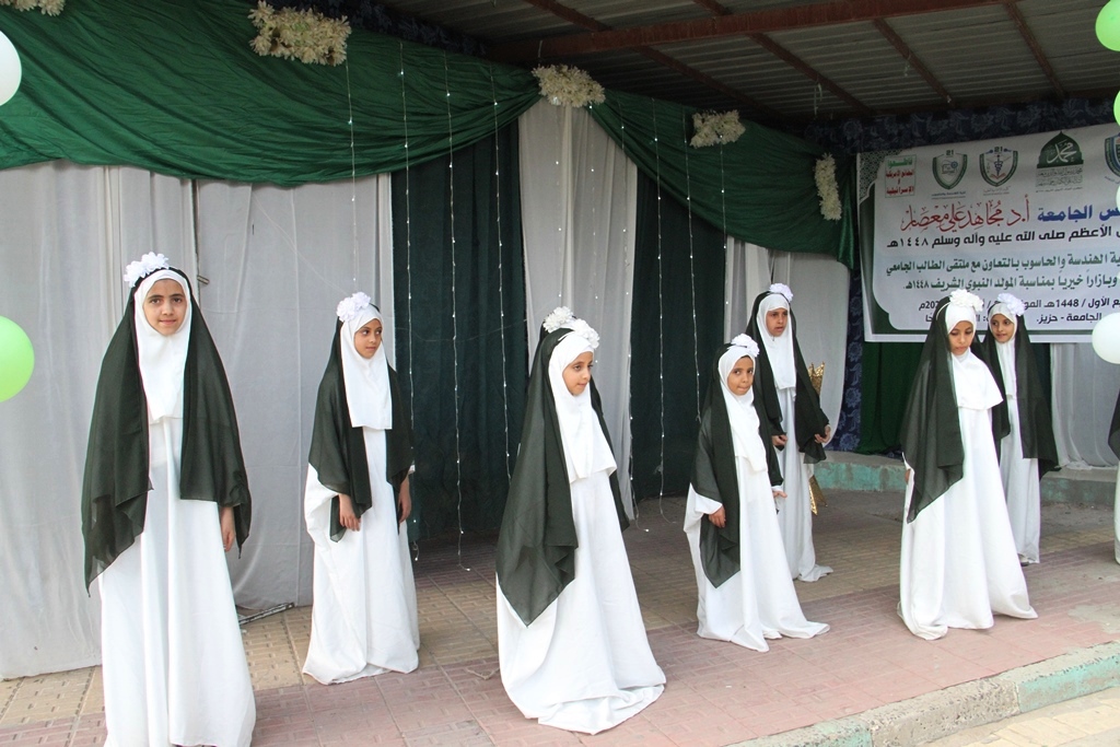 Eight girls in white dresses with dark green head coverings perform on an outdoor stage with a green backdrop and a banner in the background.