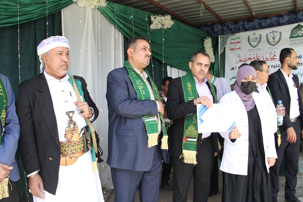 Group of formally dressed people on a stage at a ceremony; a man wears a ceremonial dagger belt while others wear green sashes; a woman in a hijab holds a certificate.