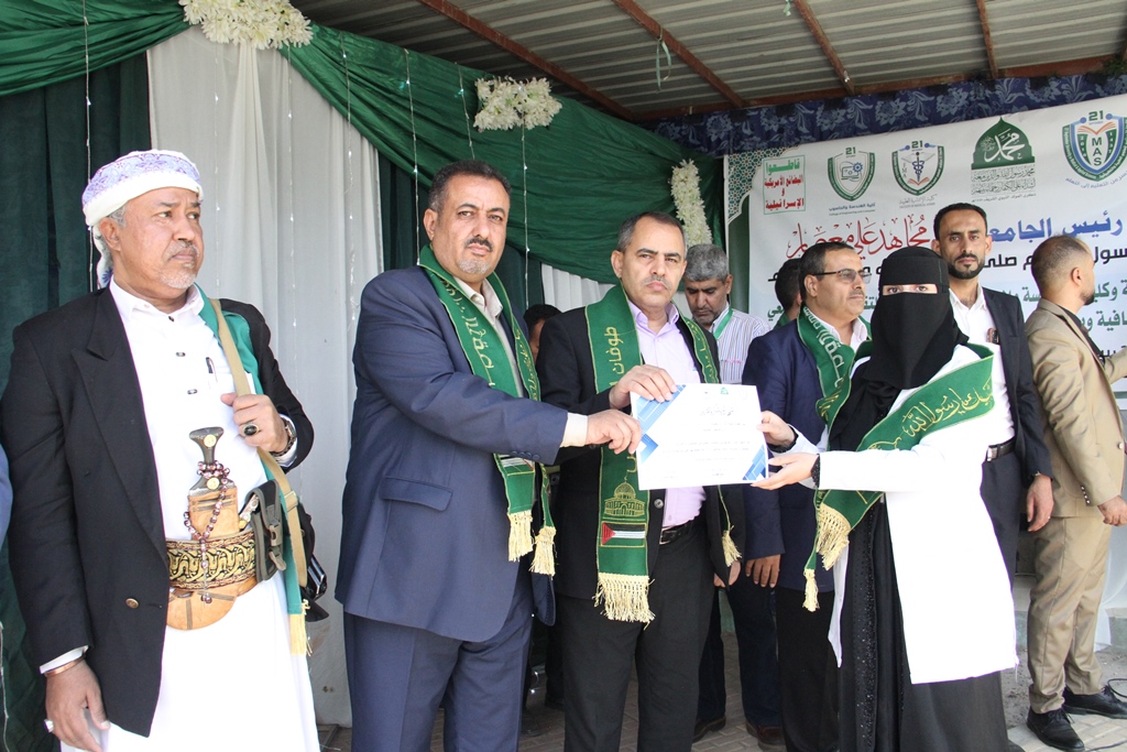Group of men in suits and traditional dress on stage presenting a certificate to a veiled recipient wearing a green sash and scarf, during an award ceremony in a decorated tent.
