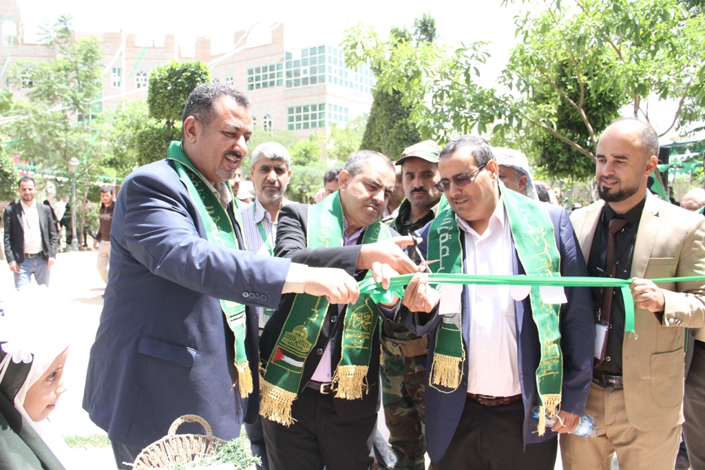 Group of men in suits cutting a green ribbon at an outdoor ceremony, with onlookers and greenery in the background.