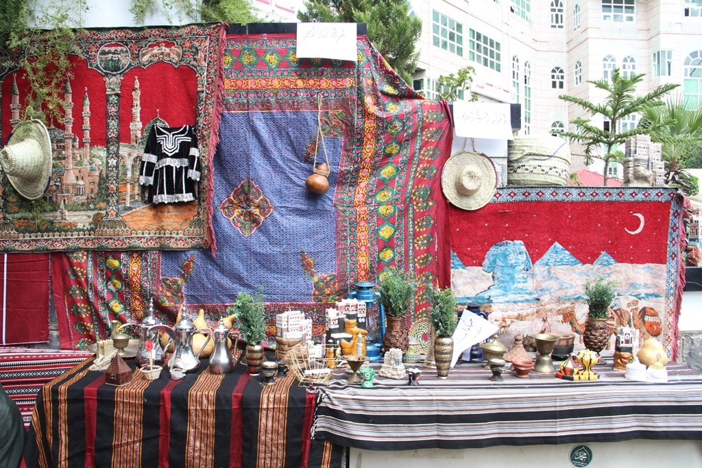 Outdoor market stall displaying vibrant carpets, textiles, and assorted pottery on striped tables.