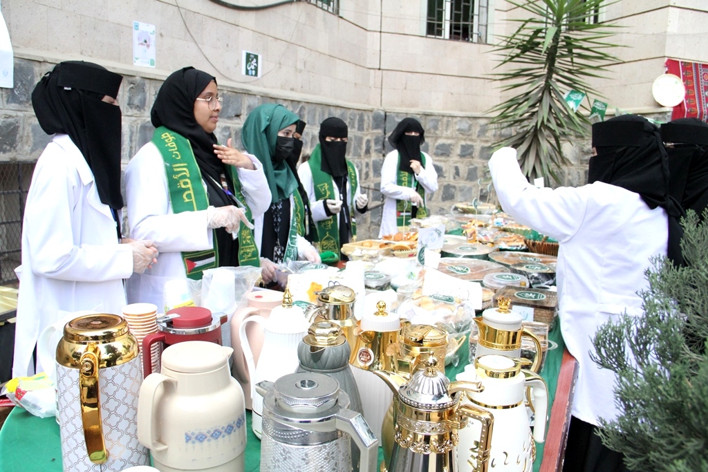 Group of women in white coats and black hijabs with green sashes stand behind a table of tea pots, kettles, and pastries at an outdoor market.