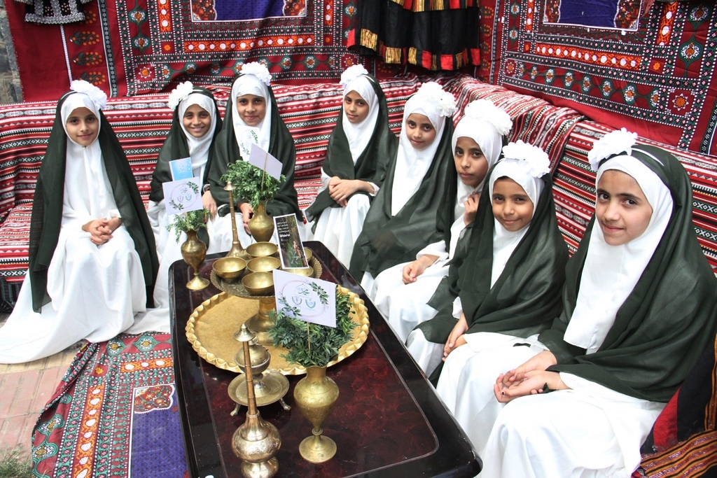 Group of young girls in white robes and dark green cloaks sit around a brass tea set on a patterned rug, smiling.