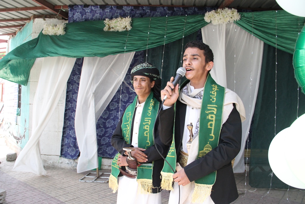 Two men in traditional robes on a decorated stage; one speaks into a microphone while the other stands with a sword, wearing green sashes with gold embroidery.