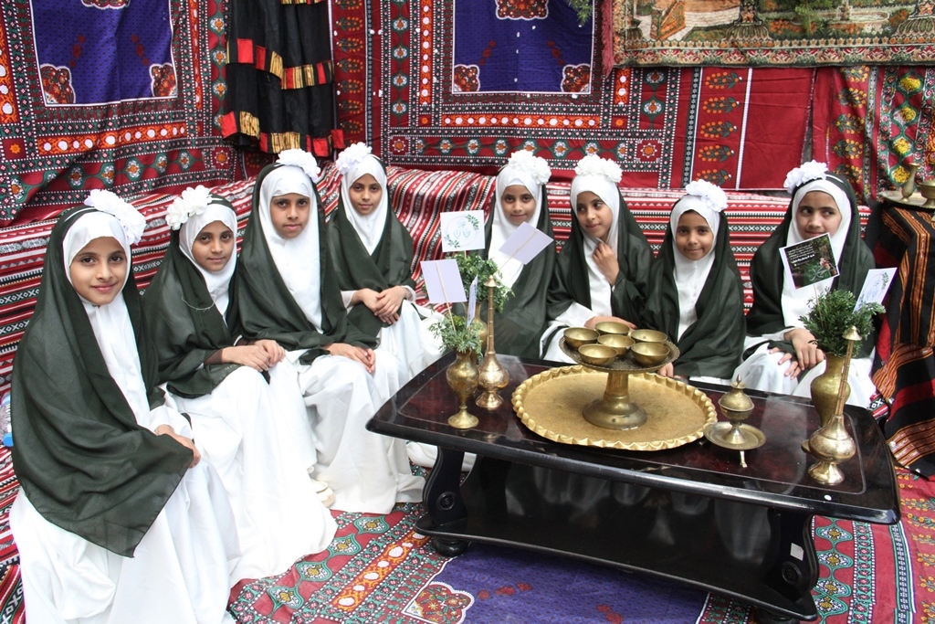 Group of young girls in white robes and green headscarves seated on a colorful patterned rug around a low table with brass dishes and decor.