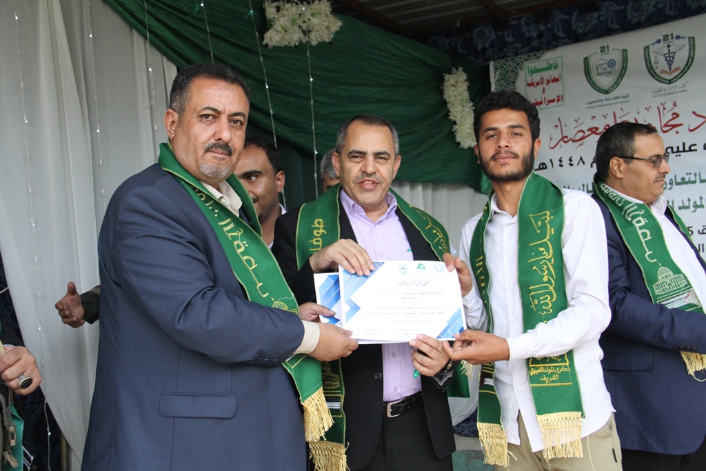 Center man in a suit hands a certificate to a recipient, flanked by others wearing green scarves at a formal award ceremony in a decorated venue.