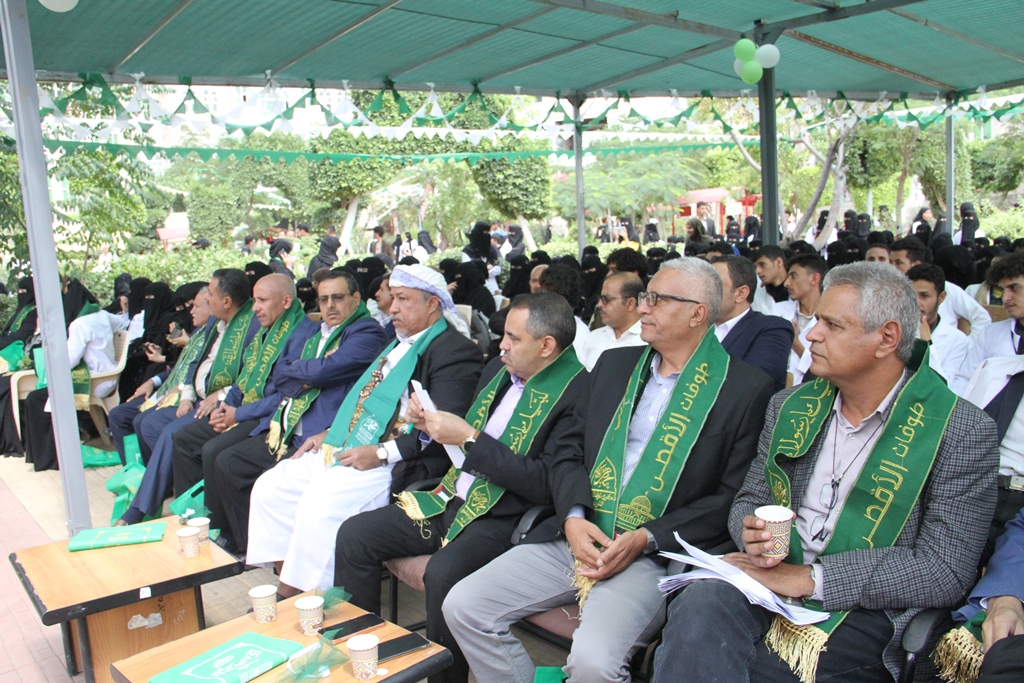 Audience seated under a green canopy at an outdoor ceremony; men in the front row wear green sashes, with women in black abayas in the background.
