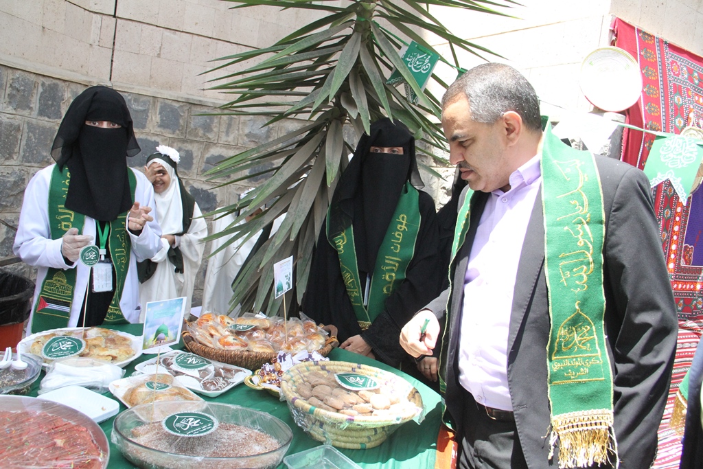 People wearing green sashes stand behind a table of pastries at an outdoor charity stall. A man in a suit browses the treats while others help."