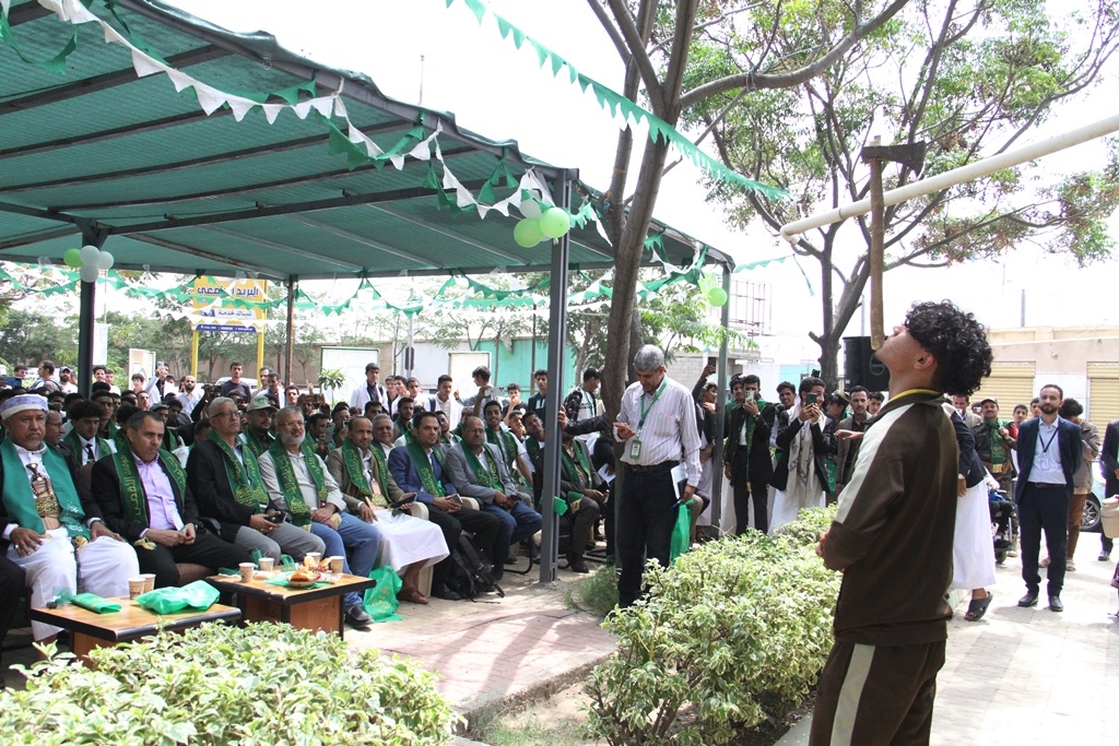 Outdoor ceremony with a large seated audience under a green canopy; attendees wear green sashes, with balloons and bunting overhead.