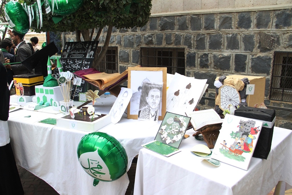 A table covered with a white cloth displays framed drawings, models, and crafts at an outdoor exhibit, with green balloons nearby.