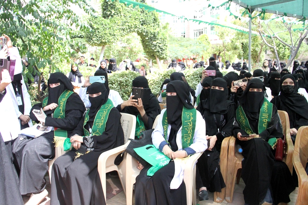 Group of women in black abayas and niqabs seated outdoors at an event, wearing green sashes with Arabic text.