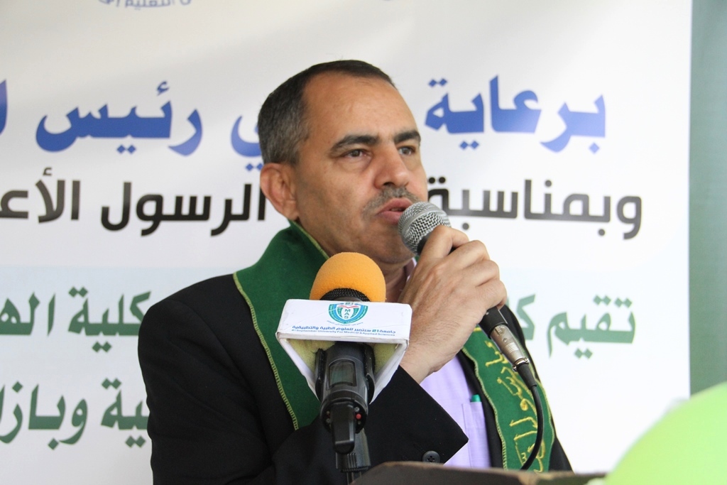 Man in a dark suit with a green stole speaks into a microphone at a podium, with an Arabic banner behind him.