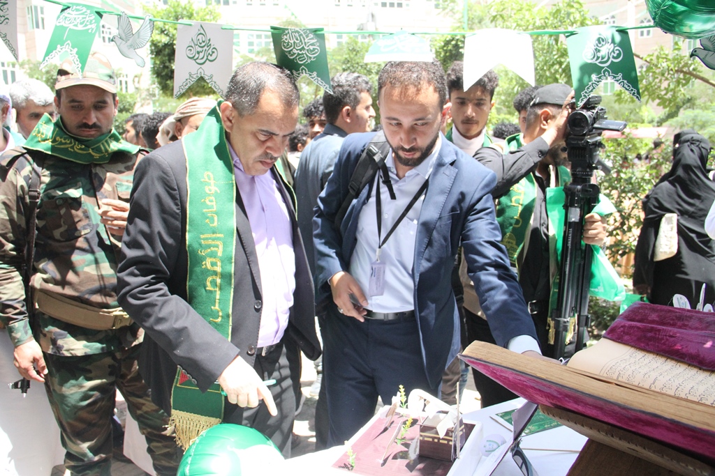 Group of men at an outdoor event around a table with an open book, green banners, and a camera crew in the background.