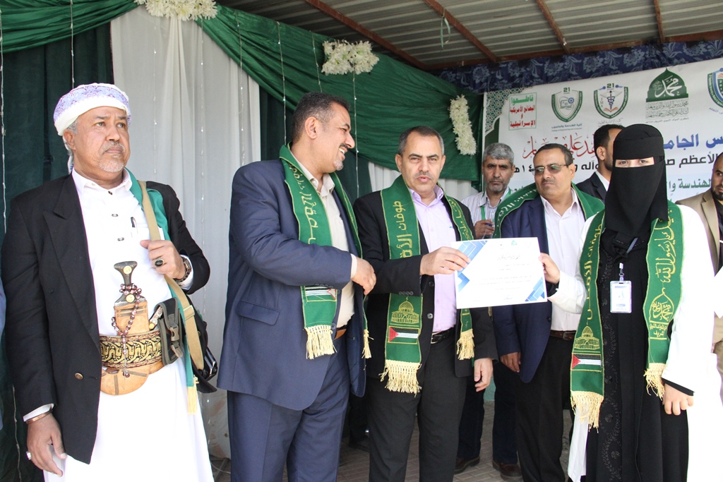 Group of men and a veiled woman on a stage, one handing a certificate to another during a ceremony with green sashes and banners in the background.