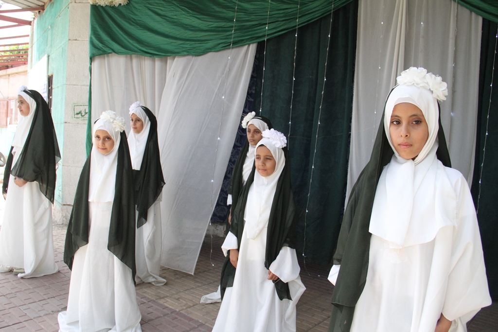 Group of girls in white robes and dark green head coverings standing outdoors in front of draped curtains with string lights.