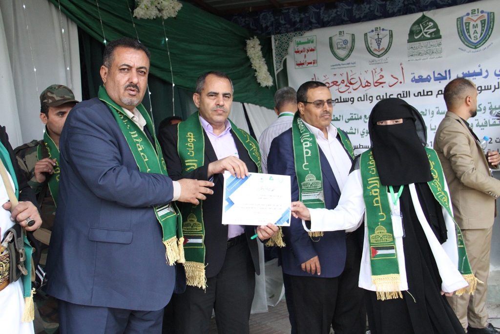 Group of officials on a stage exchanging a certificate during a formal event, with green scarves and a banner in Arabic in the background.