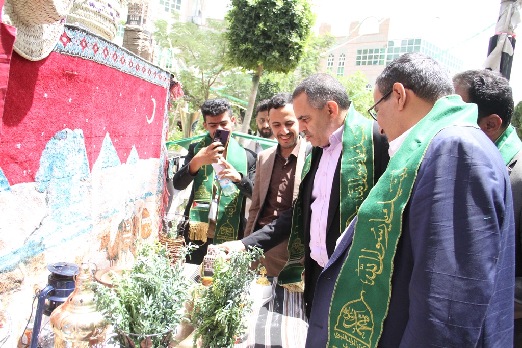 Group of men in green ceremonial sashes examining a display of plants and jars at an outdoor event, one man capturing a photo with his phone.