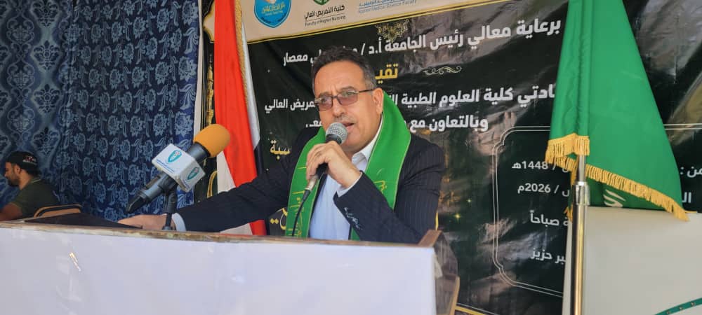 Man in a suit with a green scarf speaks into a microphone at a podium, with a black banner and green flag behind him.