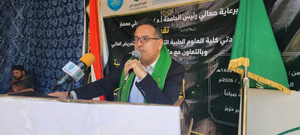 Man at a podium speaking into a microphone with a green scarf, banners and flags in the background (likely a formal event).