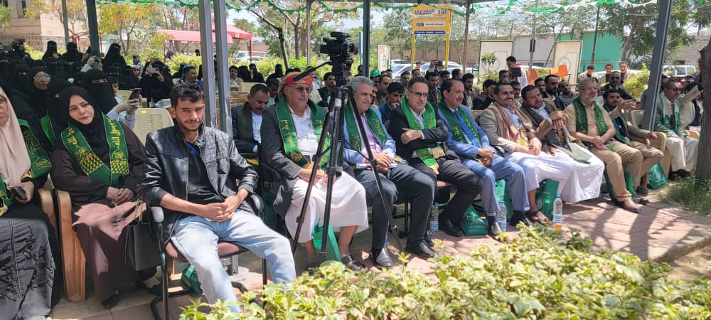 Audience seated outdoors at a ceremony; several people wear green sashes, with a camera on a tripod visible at the front.