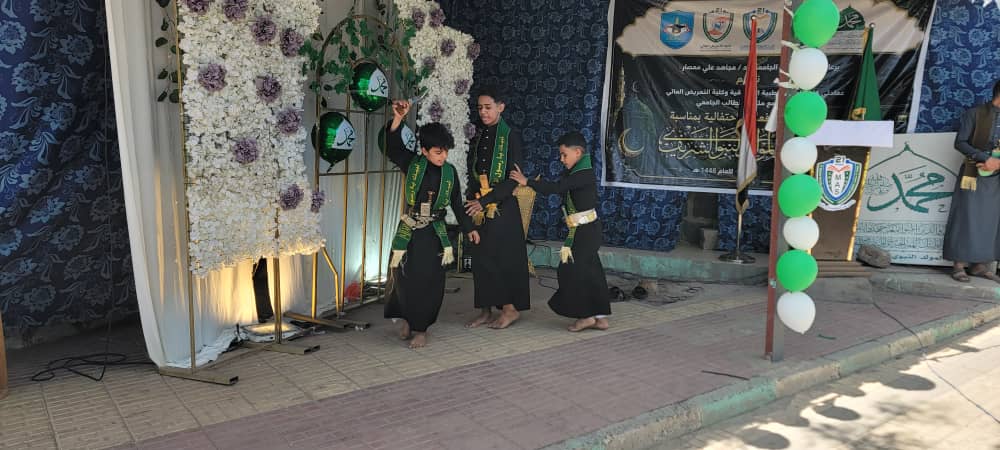 Three boys in black robes with green sashes perform a traditional dance on a decorated stage with flowers and balloons in the background.