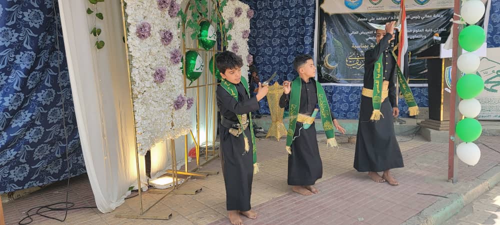 Three barefoot boys in black robes with green sashes perform a traditional dance on a stage with floral decor and a banner in the background.