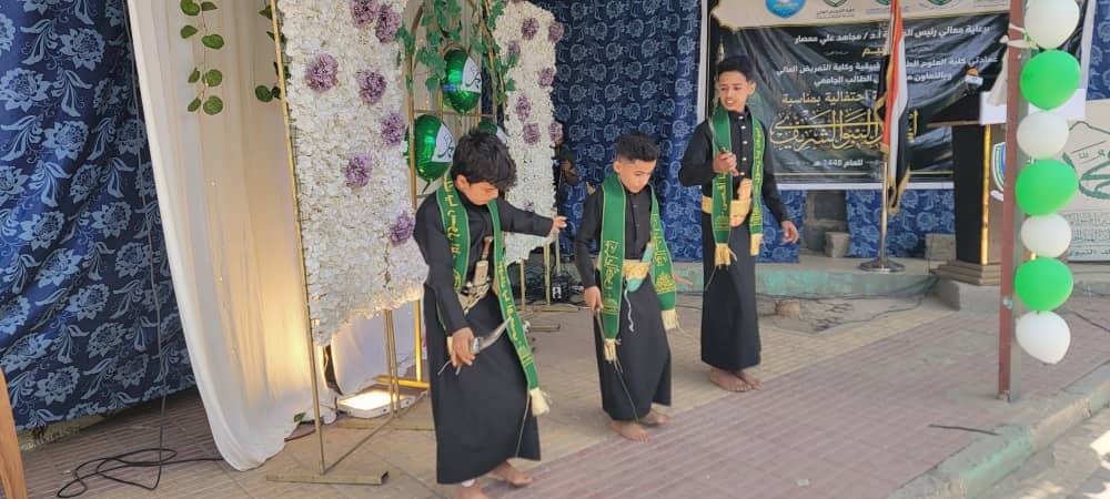 Three boys in black robes with green sashes perform a traditional dance on a sidewalk stage with a floral backdrop and banners in the background.