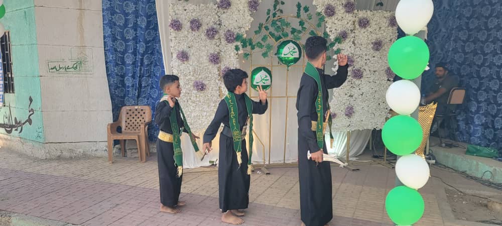 Four boys in black robes with green sashes perform a ceremonial dance on a stage decorated with a white floral backdrop and green‑white balloons.