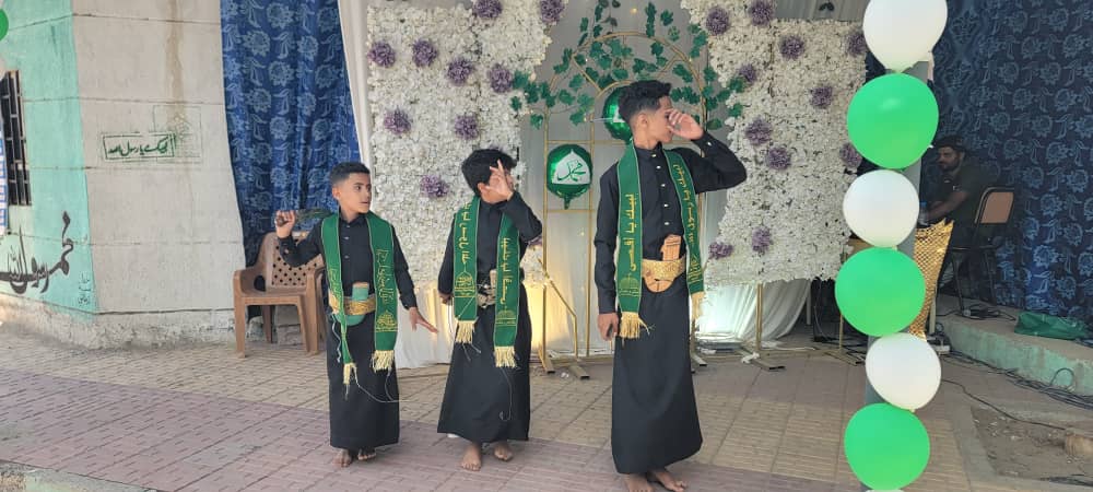 Three boys in black robes with green sashes perform on a floral stage with balloons nearby.