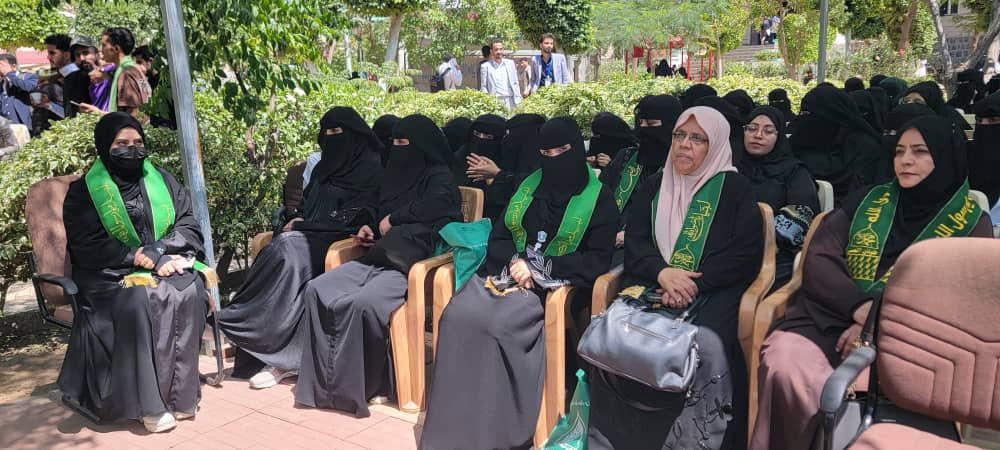 Group of women in black abayas with green sashes seated outdoors at a ceremony.