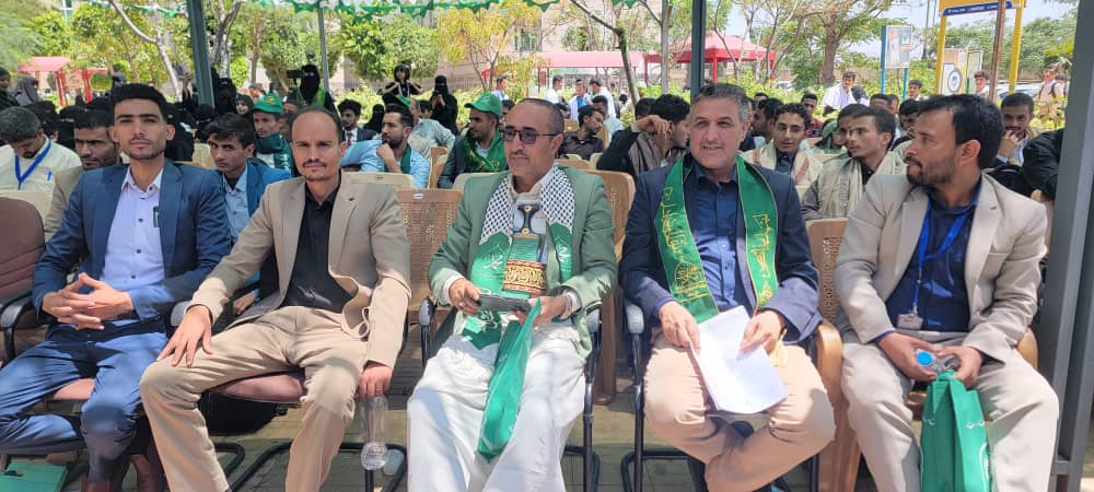 Row of men in suits seated at an outdoor ceremony, several wearing green ceremonial sashes as attendees watch.