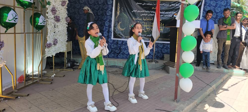 Two young girls in green skirts sing into microphones on a small stage at an outdoor celebration, with green/white balloons and a crowd behind them.