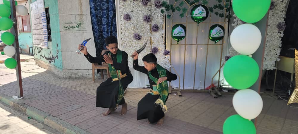 Two young men in black robes kneel on a sidewalk, wielding curved swords as part of a ceremonial performance beside a flower-decorated backdrop and green-and-white balloons.
