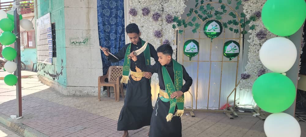 Two boys in black traditional robes with green sashes perform a dance on a sidewalk stage with a flower backdrop and green/white balloons.