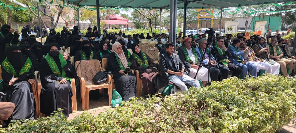 Audience gathered under a tent for a formal ceremony; many attendees wear green sashes, women in hijabs and abayas sit in rows.