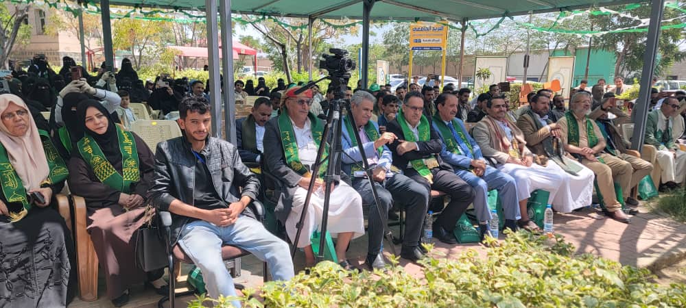 Group of people seated under a green tent at an outdoor ceremony, some wearing green sashes; a camera on a tripod in front of them.