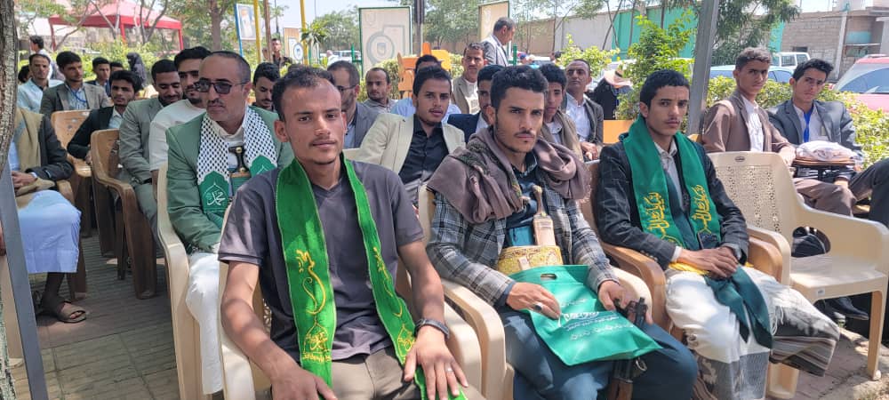 Group of men seated in plastic chairs at an outdoor ceremony; several wear green sashes.