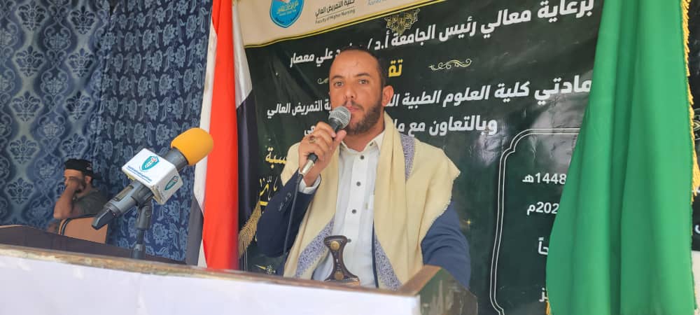 Man with a scarf speaks into a microphone at a podium during a formal event, with a green banner and flag behind him.