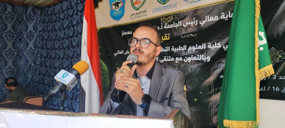 Man in a gray suit speaks into a microphone at a podium during a formal event, with Arabic banners and flags in the background.