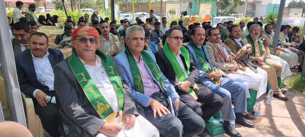 Row of men in suits seated outdoors at a ceremony, many wearing green sashes with gold text.