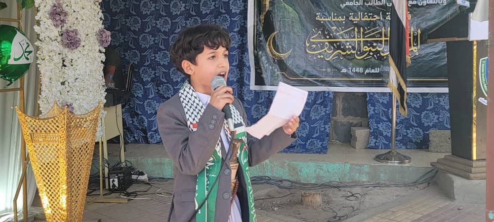 Young boy in a gray suit and green scarf speaks into a microphone at a ceremony on a decorated stage.