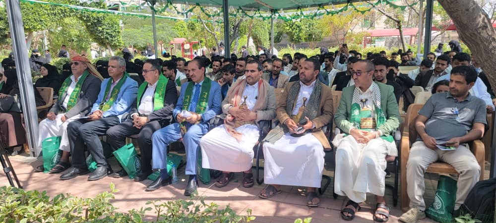Group of attendees seated under a canopy at an outdoor formal event, many wearing green sashes and holding programs? bags nearby.