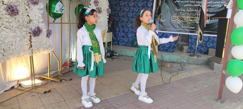 Two young girls in green and white outfits sing into a microphone on a decorated stage at an event.