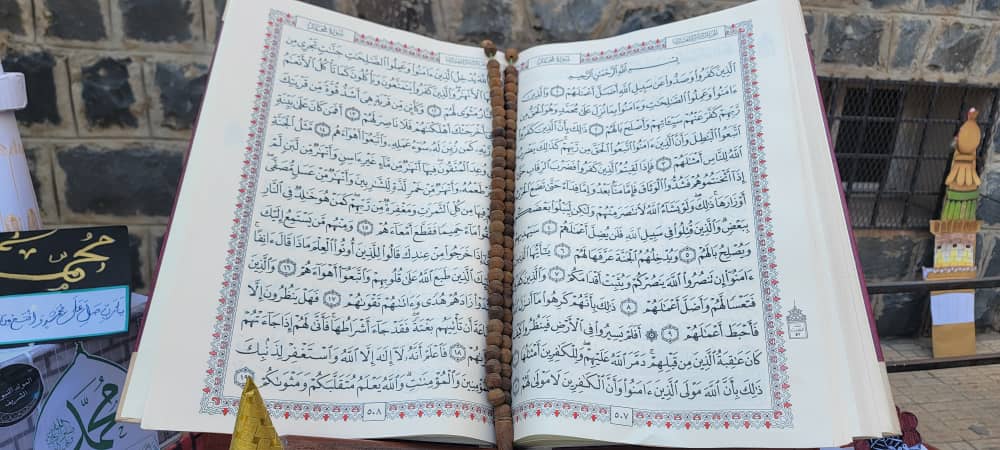 Open Quran with a string of prayer beads resting on the pages, ornate borders, displayed outdoors against a stone wall.