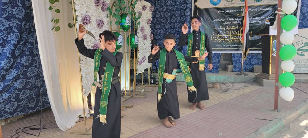 Three boys in black robes with green sashes performing a dance on a sidewalk stage under a decorated backdrop and banners in the background.