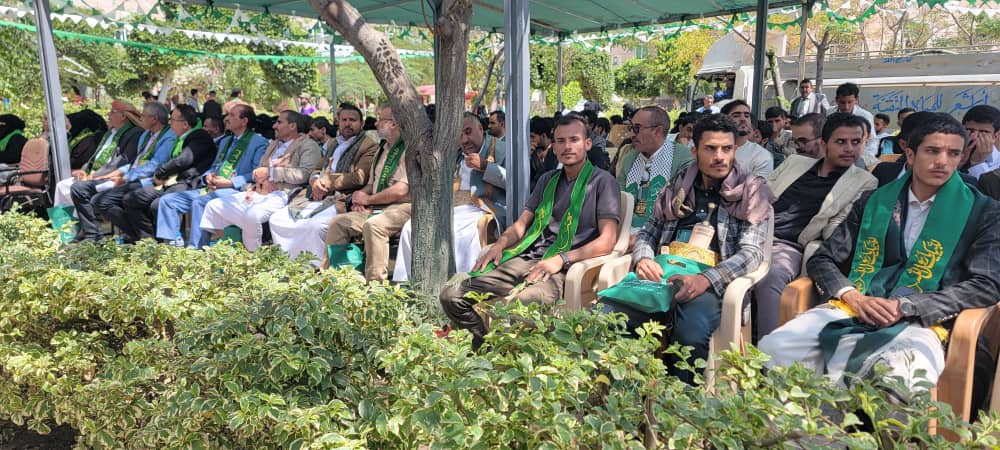 Group of men seated in rows under a canopy at an outdoor ceremony, many wearing green sashes across their chests.