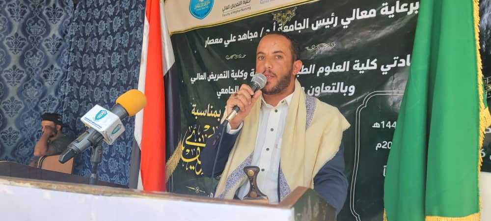 A man in a light scarf speaks into a microphone at a podium during a formal event, with Arabic banner and flags behind him.