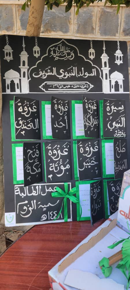 Black display board with Arabic calligraphy panels, lantern illustrations, and green ribbons for an event board on a wall.