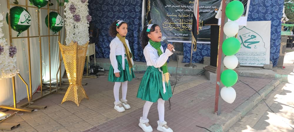 Two young girls in white blouses and green skirts perform on a stage with a microphone, flanked by green-and-white balloons and floral decor.