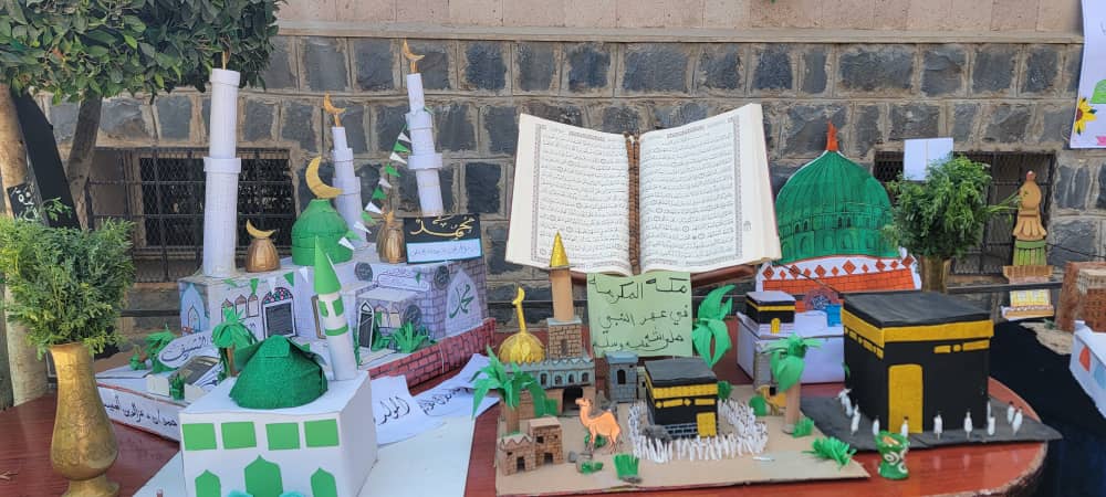 Tabletop display of handmade paper models of mosques and Islamic landmarks, centered by a large open Qur'an sculpture in the middle.